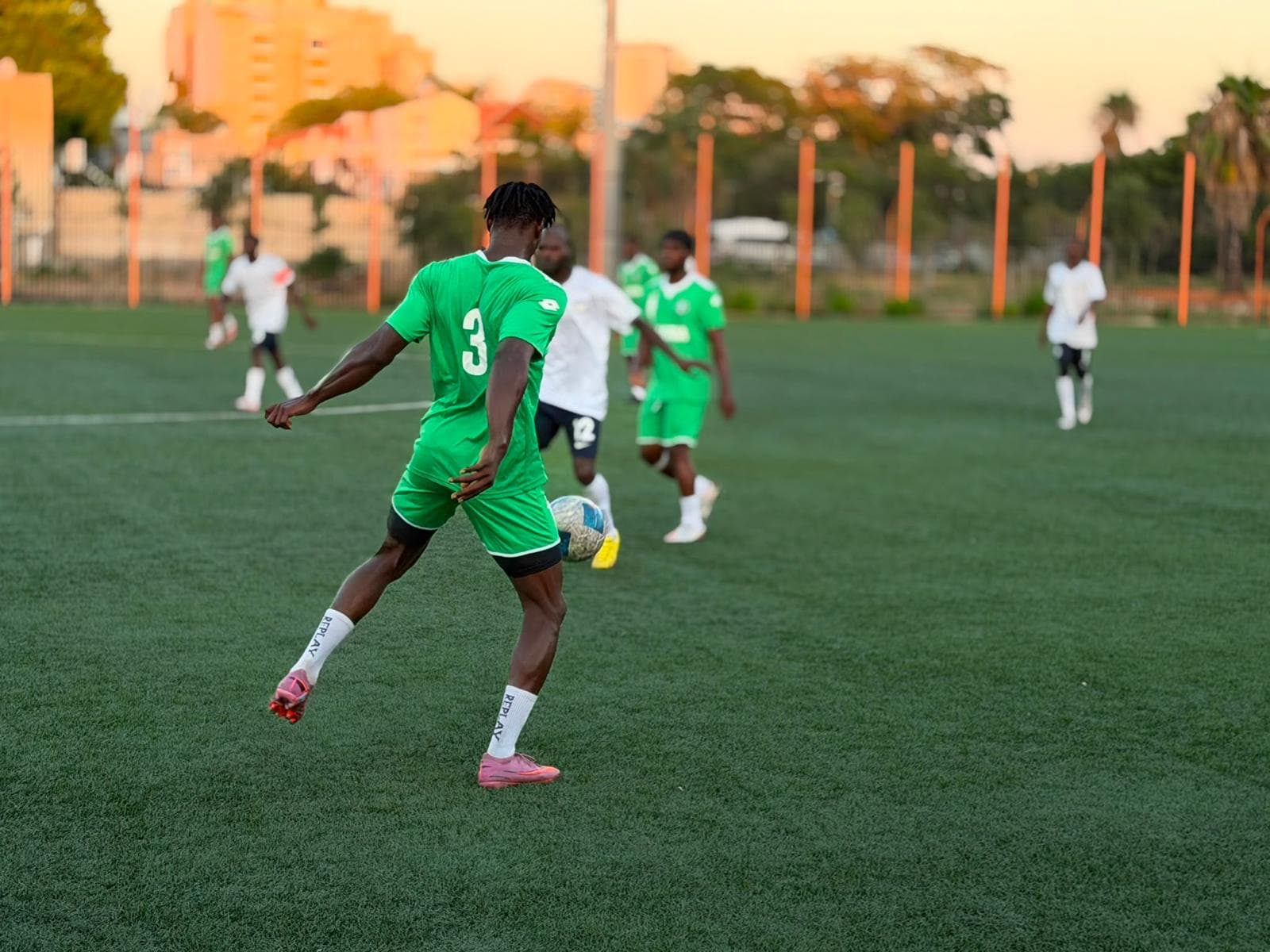 Players in green and white kits during a match on a floodlit pitch at golden hour, a city skyline behind.