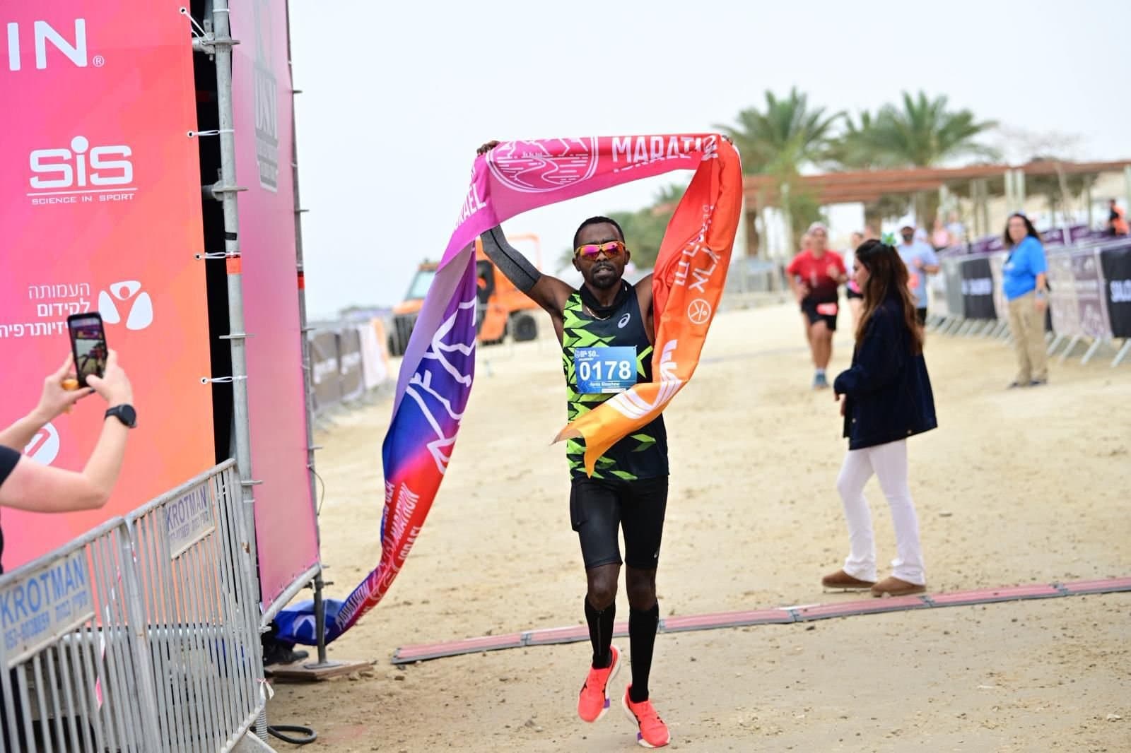 A runner breaks the finish tape on a beachfront course, arms raised.
