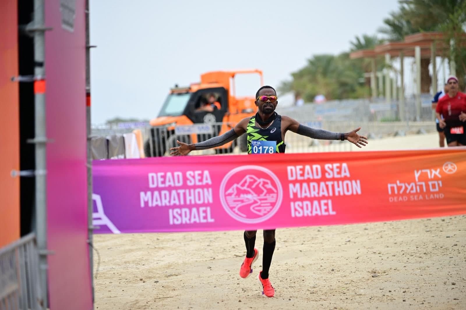 An Israeli distance runner crosses a finish line, arms wide, beneath a Dead Sea Marathon Israel banner.