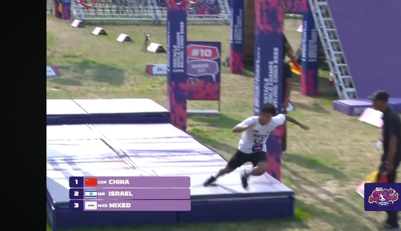 Nevo Beck races across an obstacle on the course in Beijing, the broadcast scoreboard showing Israel in second place.