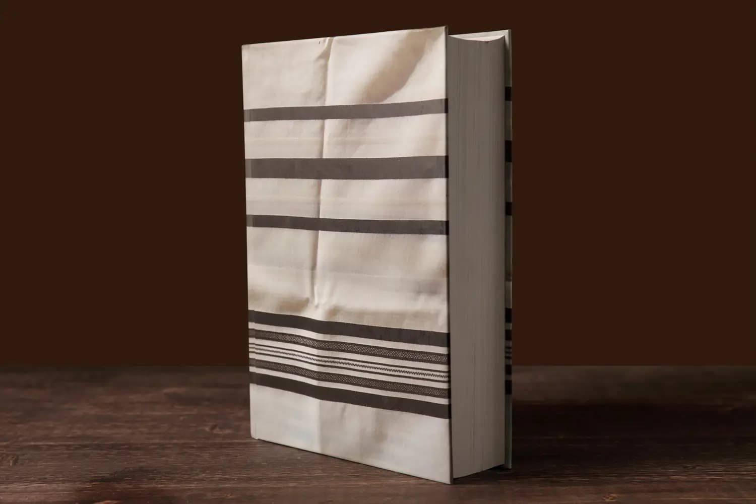 A thick book bound in a prayer-shawl cover standing on a wooden table against a dark background.