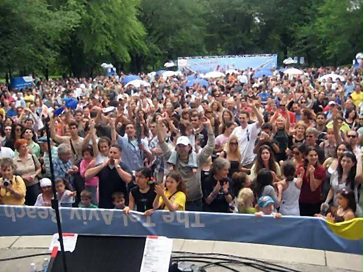 A large crowd in Central Park raising their arms and dancing in front of a banner reading The Tel Aviv Beach.