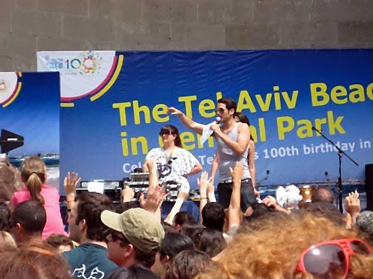 Performers on stage before a raised-arm crowd, beneath a banner reading The Tel Aviv Beach in Central Park, celebrating Tel Aviv's 100th birthday.